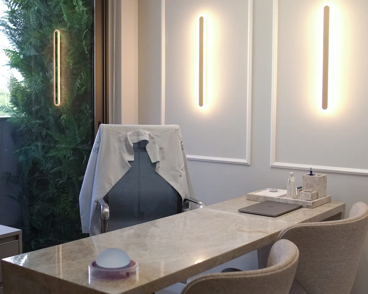 A sala de consulta: mesa de mármore bege, jaleco branco no encosto da cadeira, duas luminárias verticais acesas na parede branca e uma parede de samambaias à esquerda.