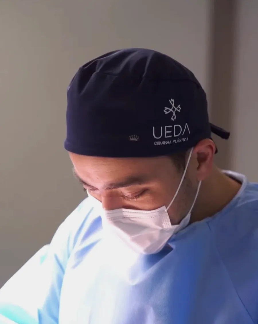Dr. Winston Ueda visto de cima, de touca com o logo Ueda e máscara, concentrado no campo cirúrgico.