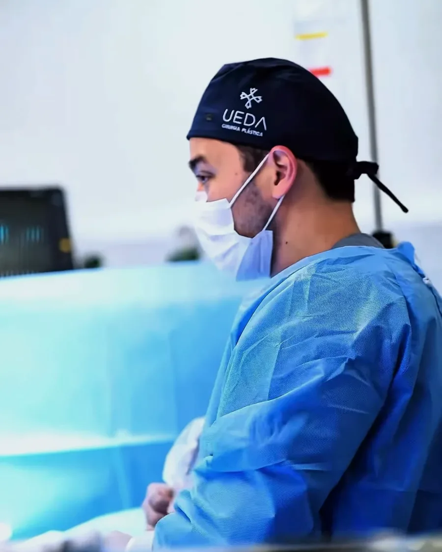 Dr. Winston Ueda de perfil no centro cirúrgico, de touca e avental azul, com o suporte de soro ao fundo.