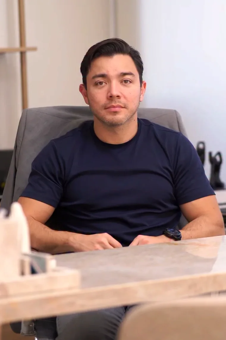 Dr. Winston Ueda sentado do outro lado da mesa de mármore do consultório, de camiseta azul-marinho, olhando para quem chega.